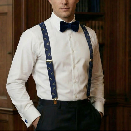 Man wearing a white dress shirt, navy bow tie, and suspenders with a wooden panel background