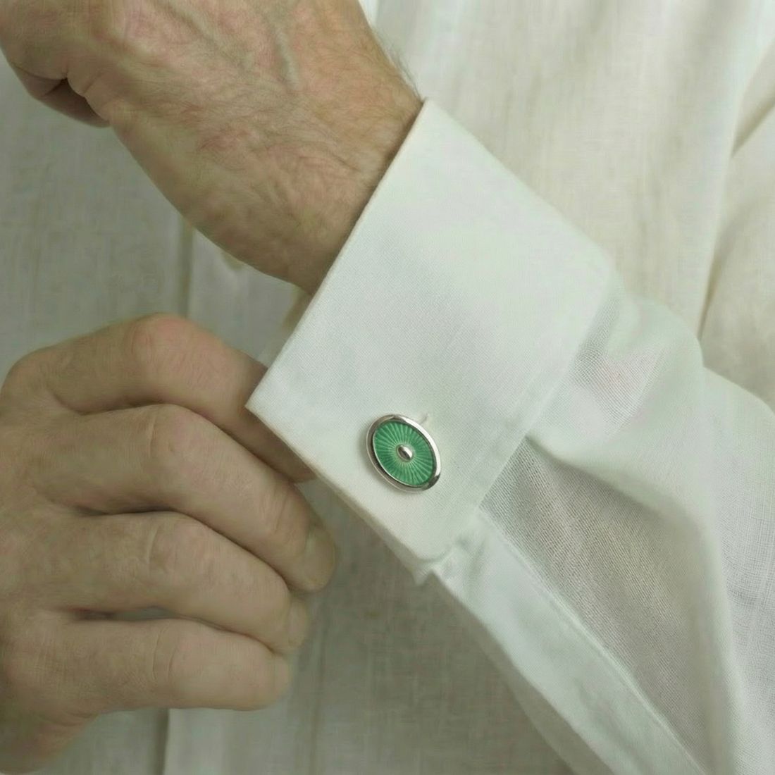 Close-up of a person adjusting a cufflink on a white shirt with a green and silver cufflink. Bown of London